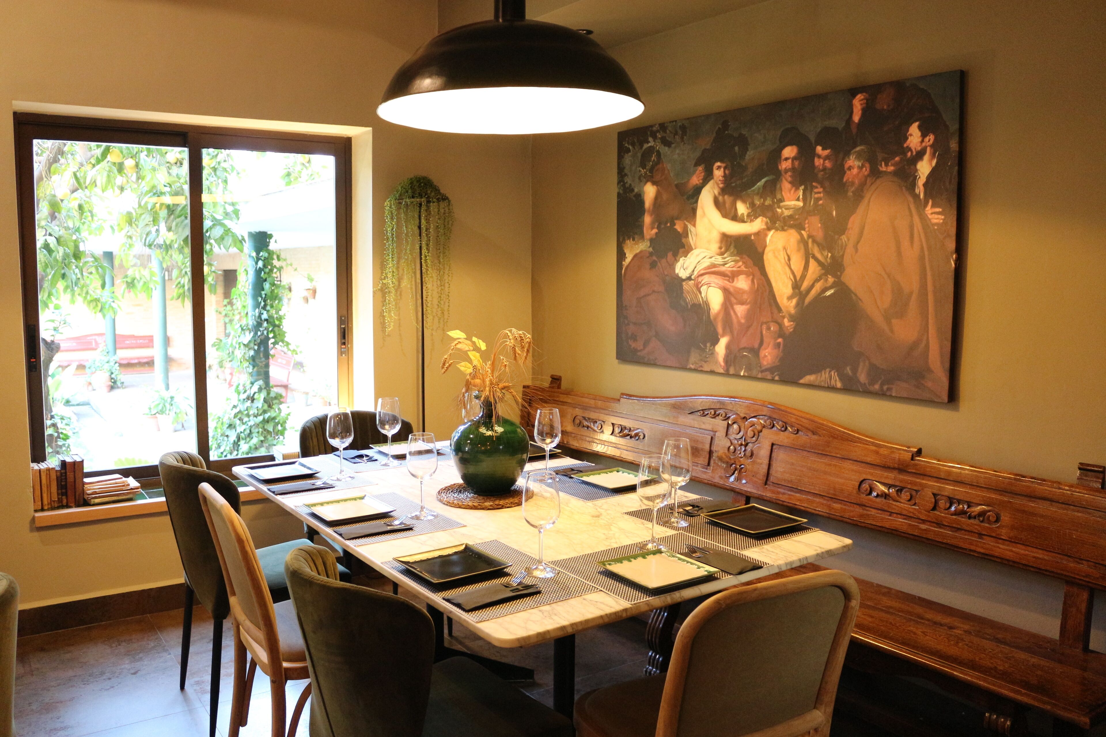 A dining area with a long table set for six, a large painting on the wall, and a window with plants.