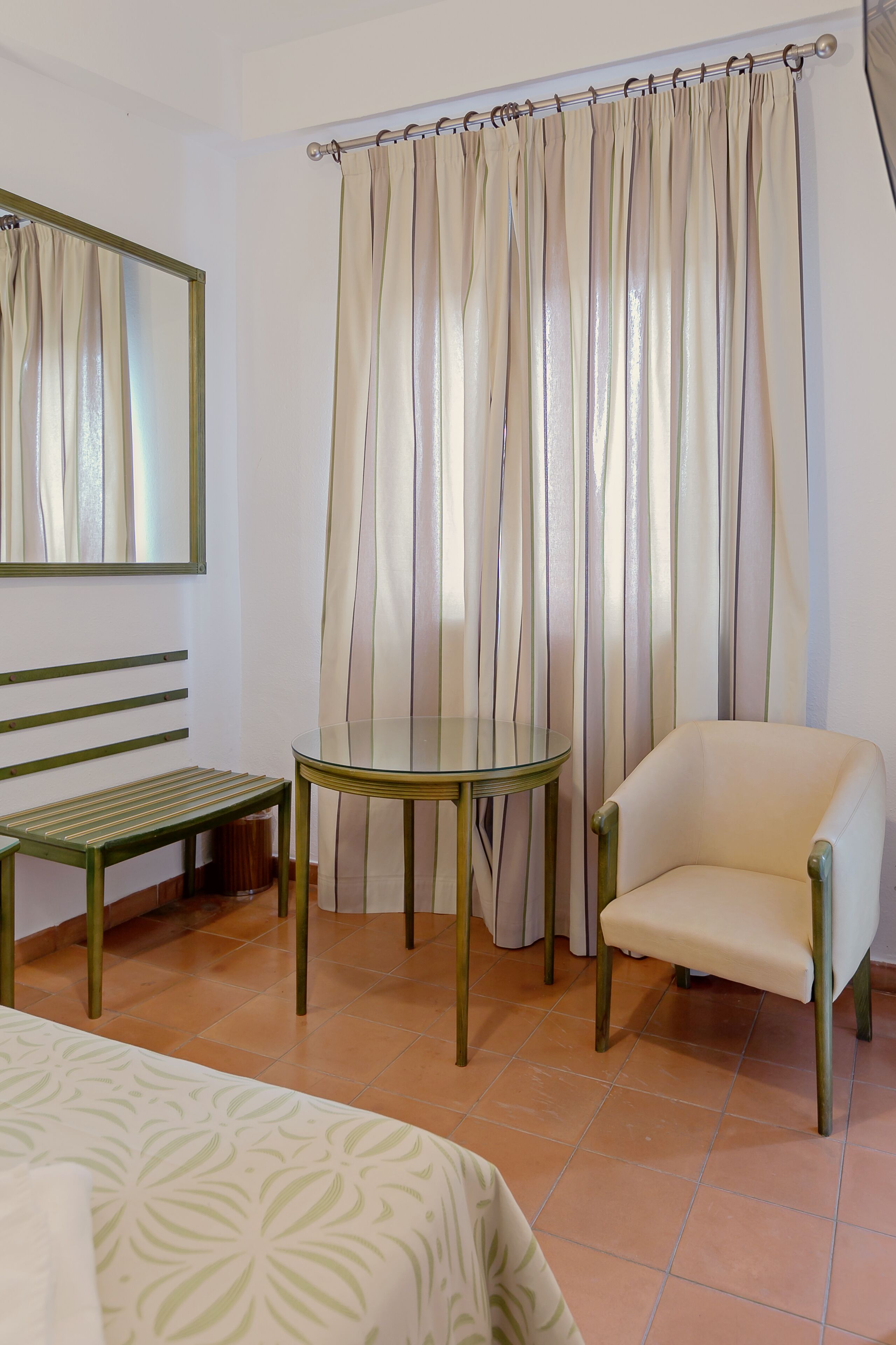 A room with a bed, a chair, a small table, a mirror, and a curtain.
