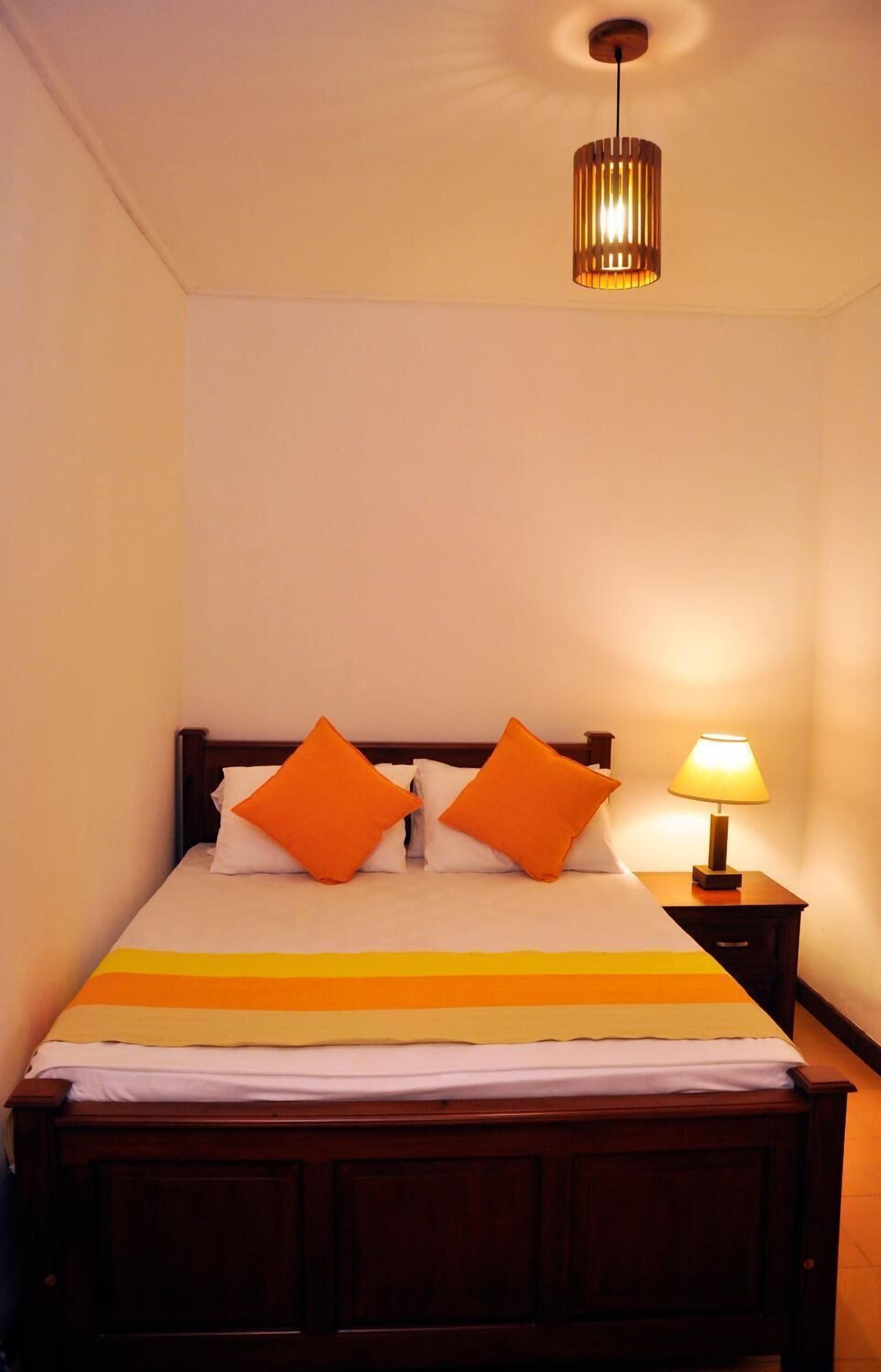 A neatly made bed with a striped bedspread, two orange pillows, a bedside lamp, and a hanging light fixture.