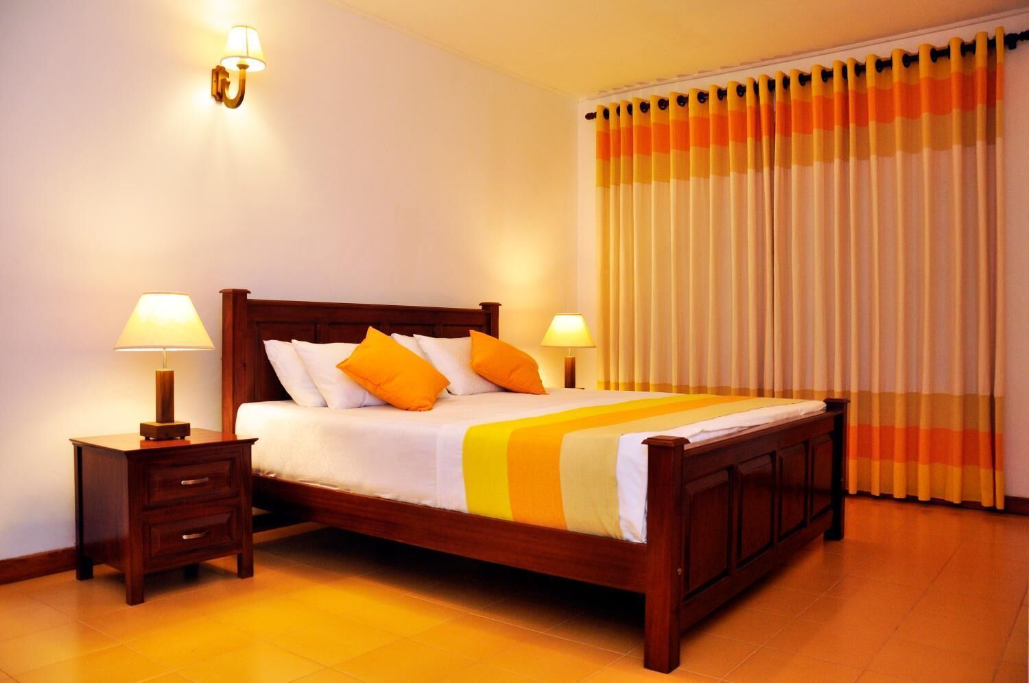 Una cama bien hecha con cabecera de madera, dos mesitas de noche con lámparas y una ventana con cortinas a rayas.