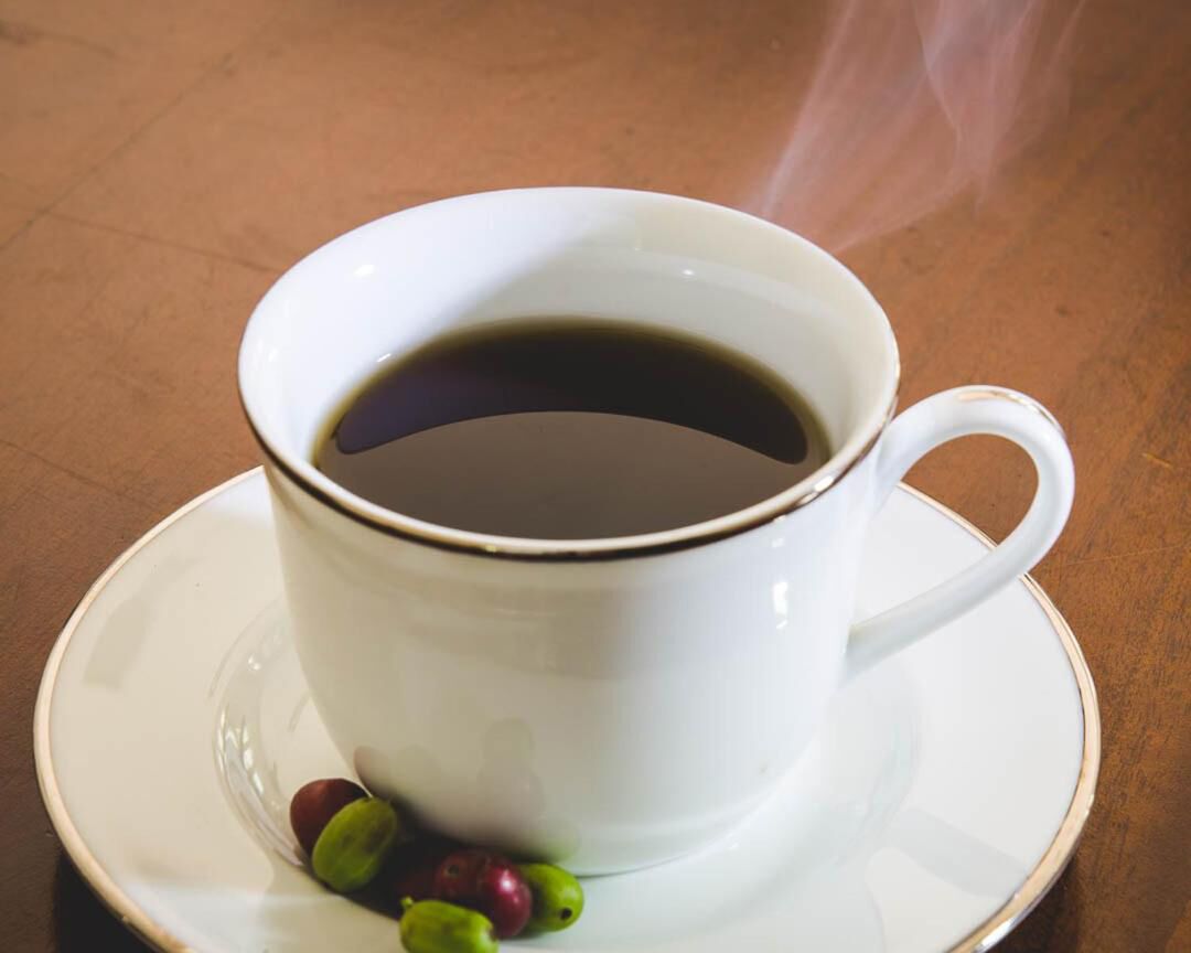 A white cup of coffee with steam on a saucer, accompanied by a few coffee beans.