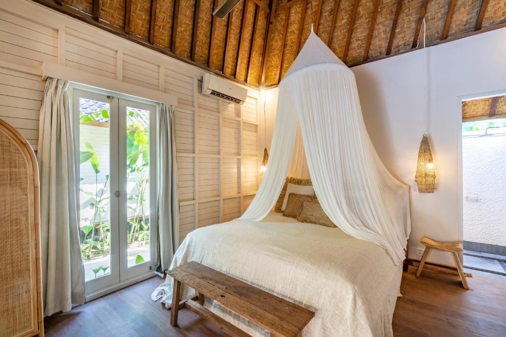 A bedroom with a canopy bed, a bench, a wicker chair, and a view of greenery through the door.