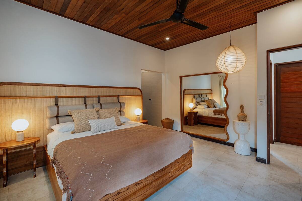 A bedroom with a large bed, wooden headboard, bedside lamps, a ceiling fan, and a mirror on the wall.