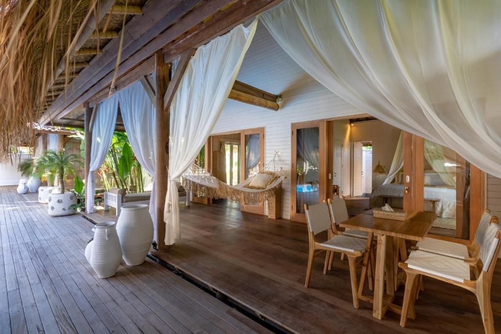 A wooden deck with a thatched roof, white curtains, a hammock, and a dining area with chairs and a table.