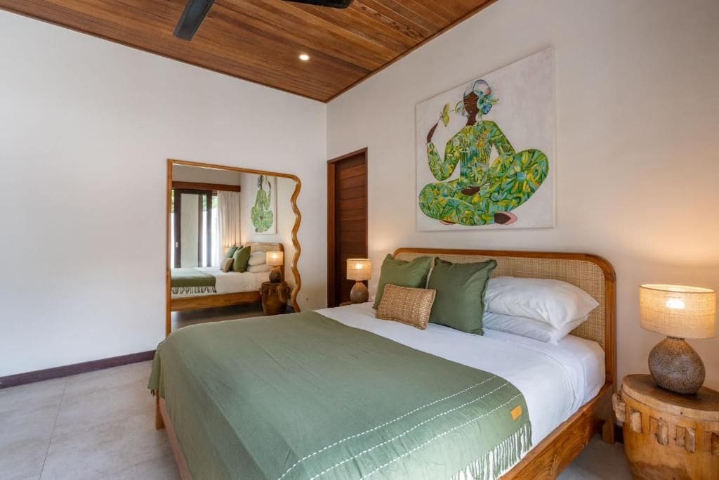 A bedroom with a wooden ceiling, a bed with green bedding, a large mirror, and a wall painting.