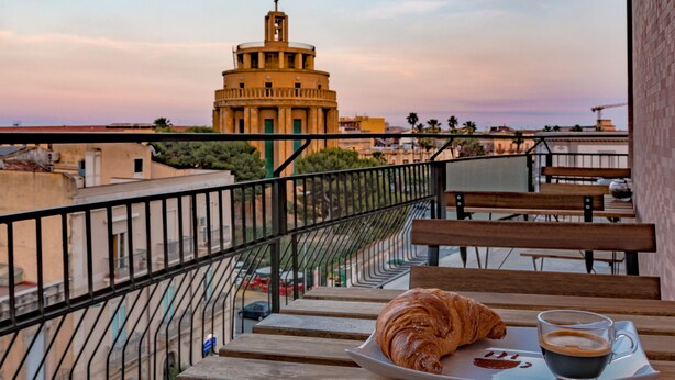 Un balcó amb un croissant i cafè en un safata, amb vista a un paisatge urbà i un edifici destacat.