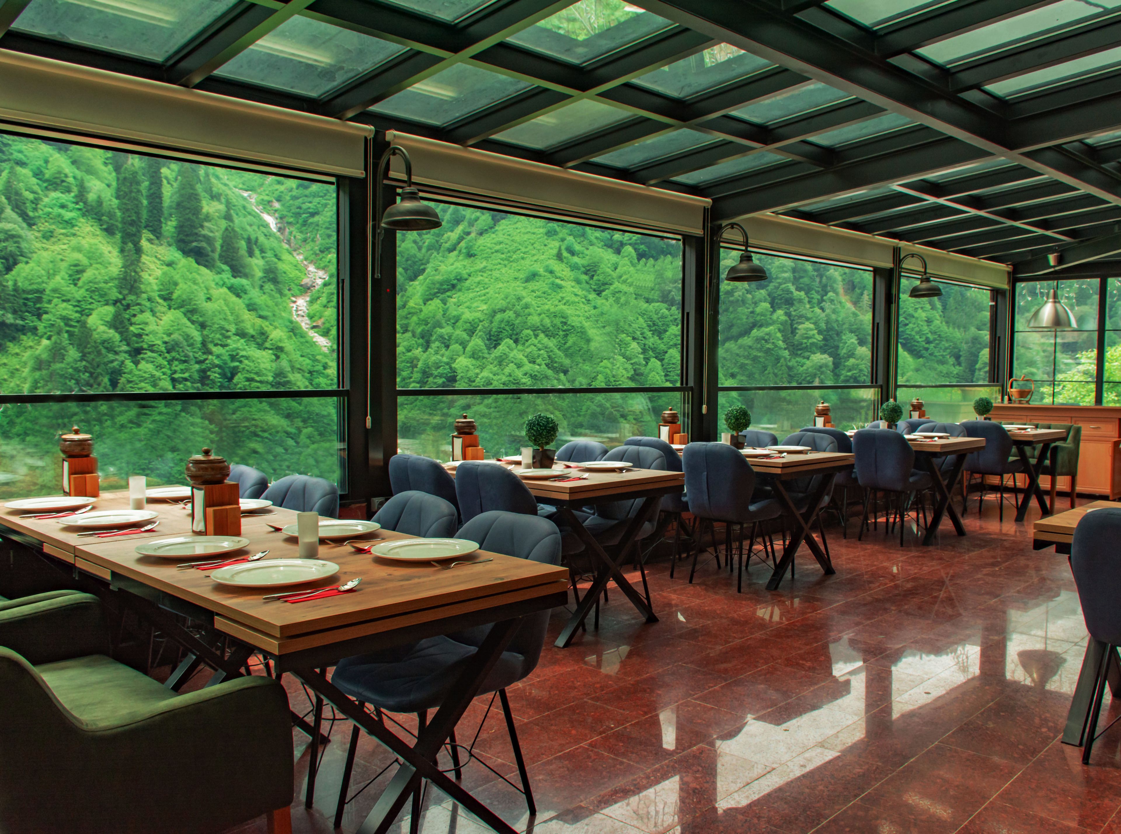 A restaurant with large windows offering a view of a forested area. Tables are set with plates, cutlery, and glasses.