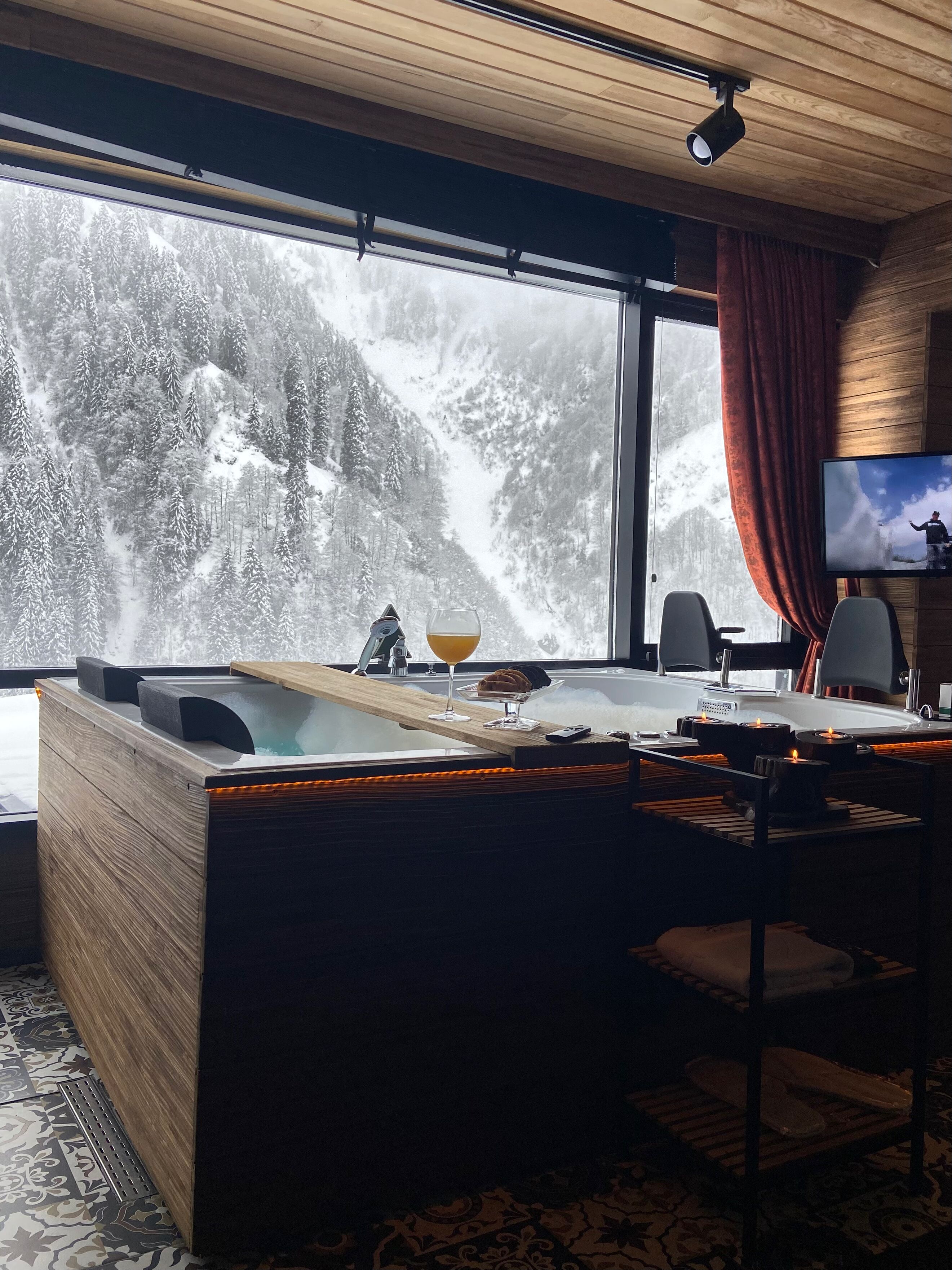 A modern room with a large window overlooking a snowy mountain. There is a wooden desk with a glass of wine and a remote control, a television on the wall, and a cozy seating area with a red curtain.