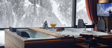 A modern room with a large window overlooking a snowy mountain. There is a wooden desk with a glass of wine and a remote control, a television on the wall, and a cozy seating area with a red curtain.