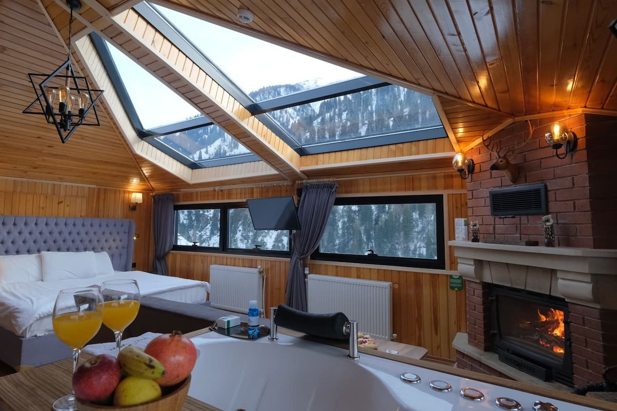 Une pièce chaleureuse avec un grand lit, une cheminée, une lucarne et un coin repas où l'on trouve des fruits et des boissons.
