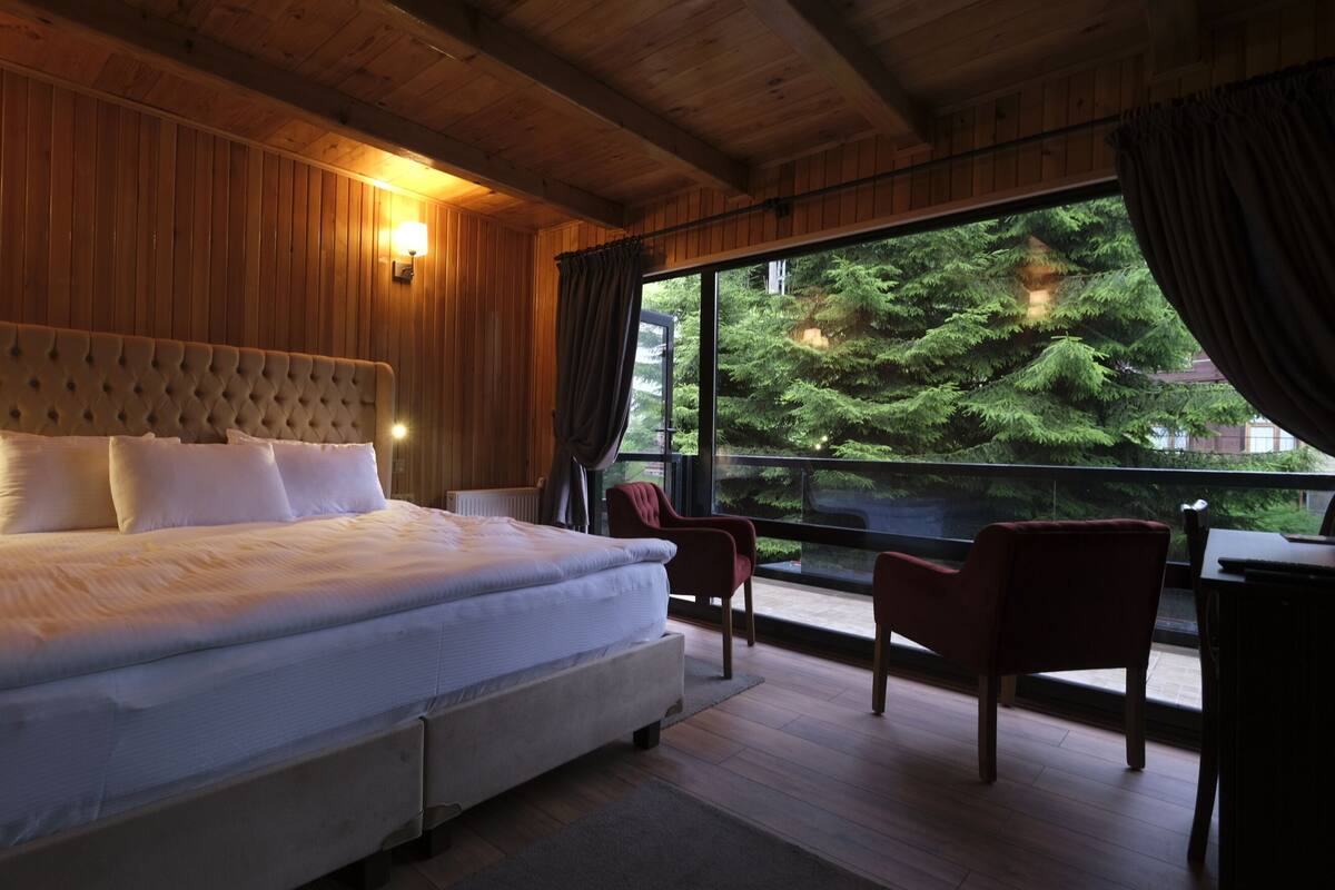 Un grand lit doté d’une tête de lit capitonnée, deux tables de chevet et une vue sur une forêt, visible à travers une grande fenêtre.