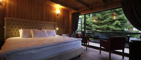 A large bed with a tufted headboard, two bedside tables, and a view of a forest through a large window.