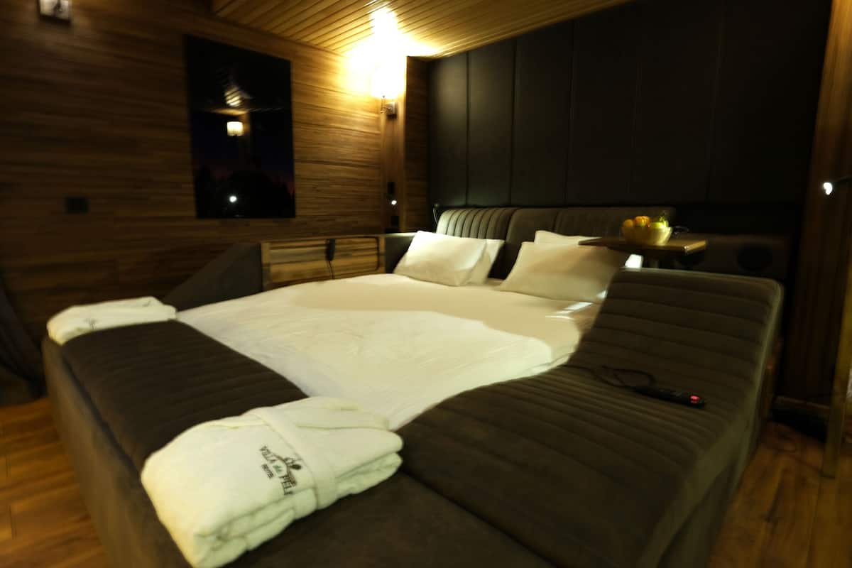 Une chambre d’hôtel moderne avec un intérieur en bois, un grand lit recouvert de draps blancs, une table de chevet avec un panier de fruits et une petite table de nuit avec une lampe.