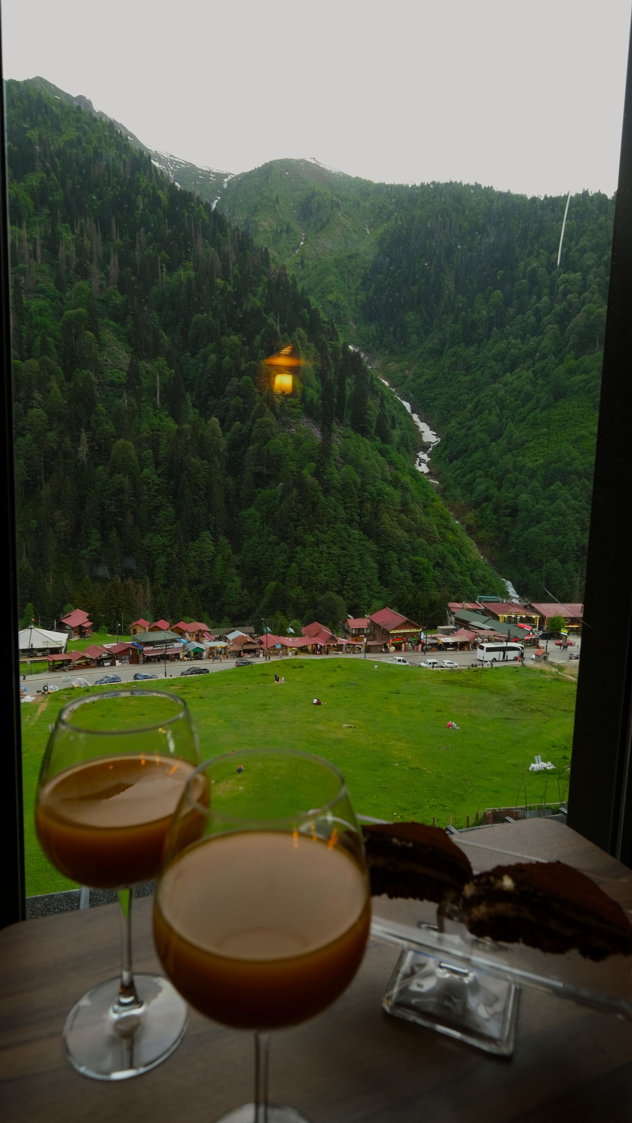 Una mesa con dos copas de vino y un postre de chocolate, con vista a un paisaje montañoso que incluye un pequeño pueblo y un río.