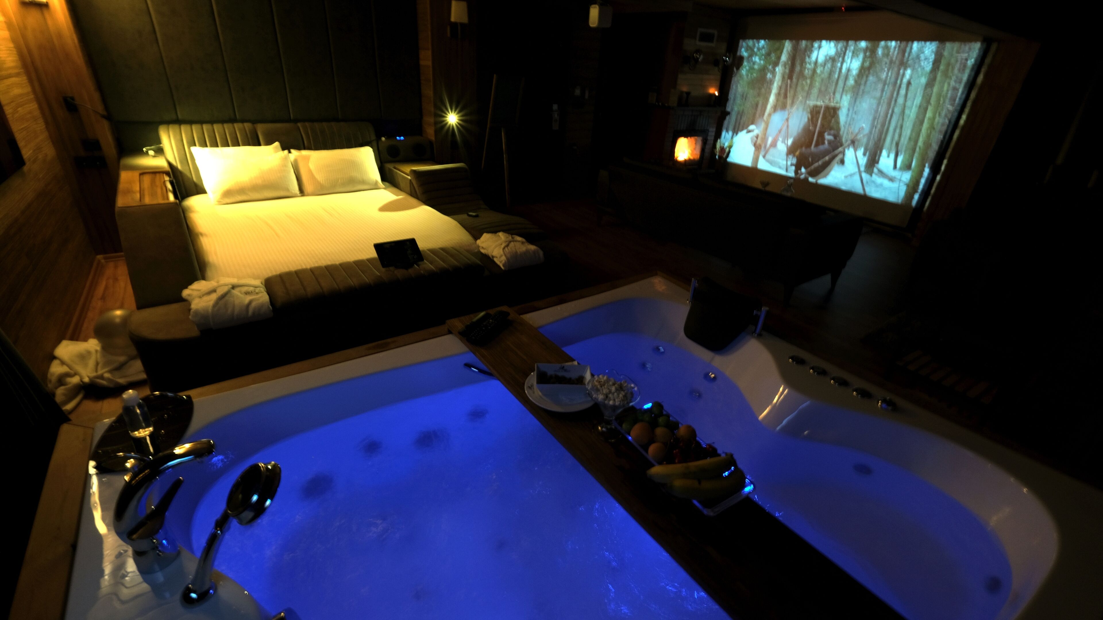 A room with a large bathtub, a bed, and a television screen displaying a forest scene.