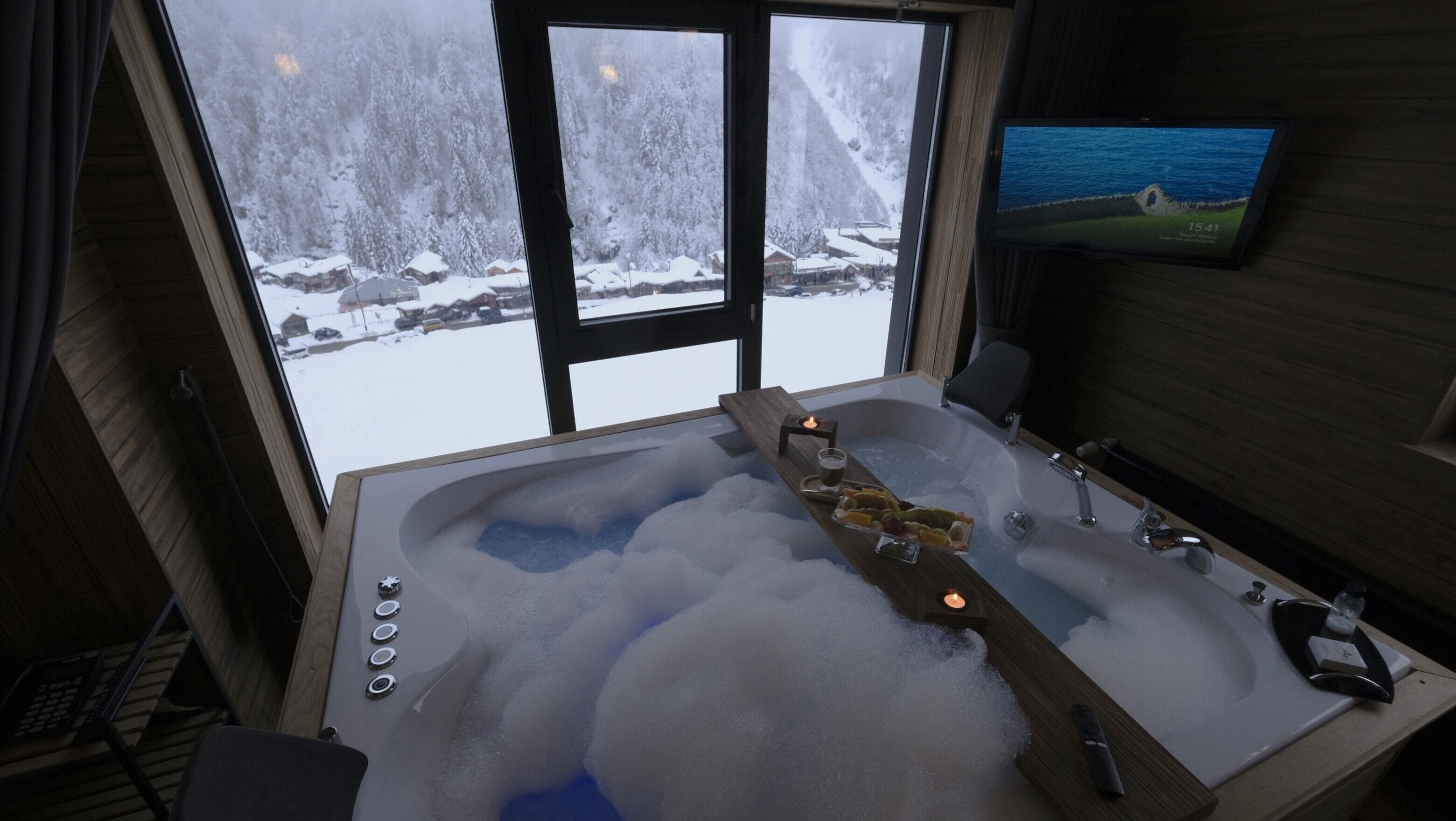 A room with a large bathtub filled with bubbles, a wooden bench, a TV on the wall, and a view of a snowy landscape through the windows.