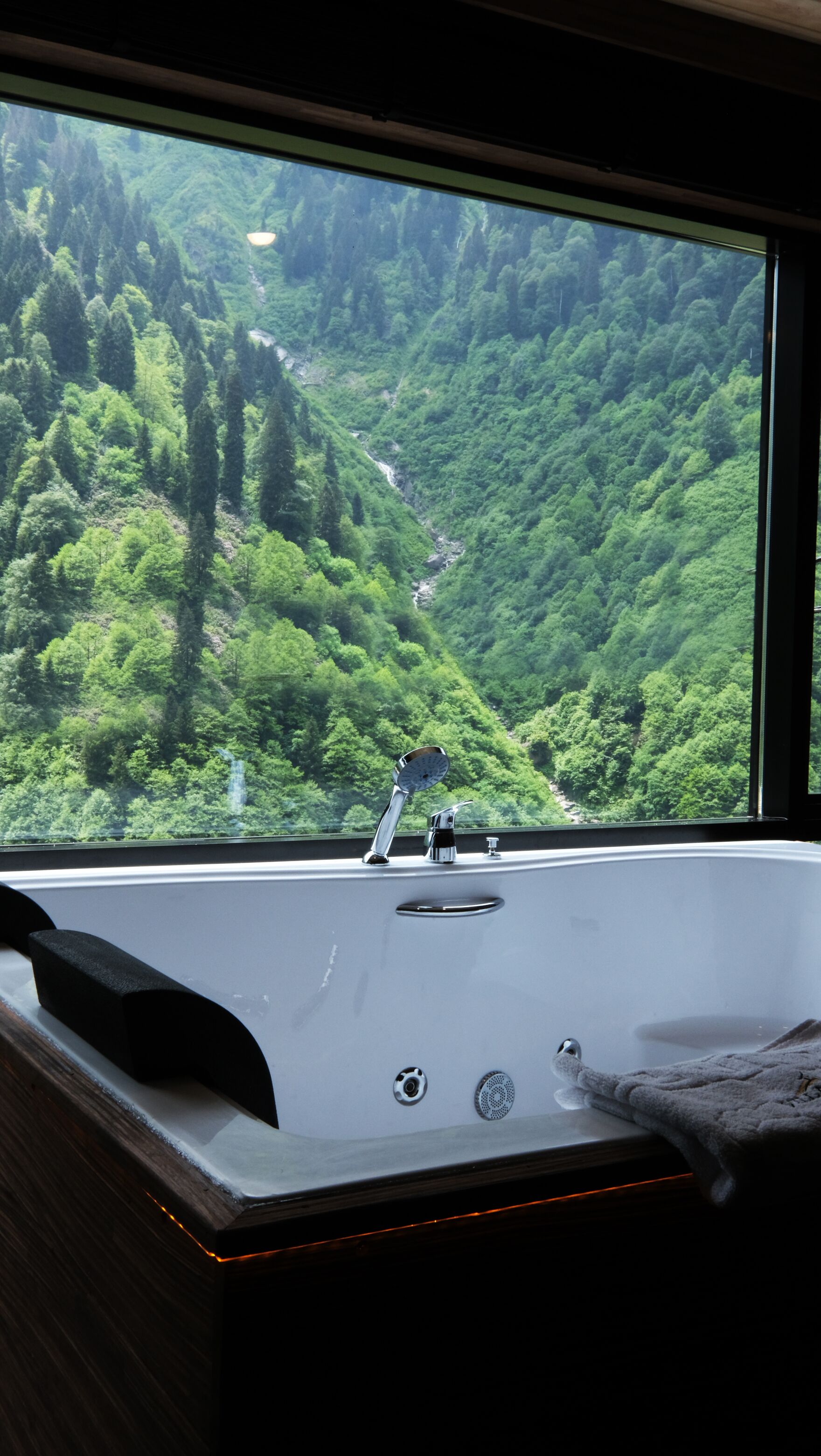 A bathroom with a bathtub, a window overlooking a forested valley, and a towel on the edge of the tub.