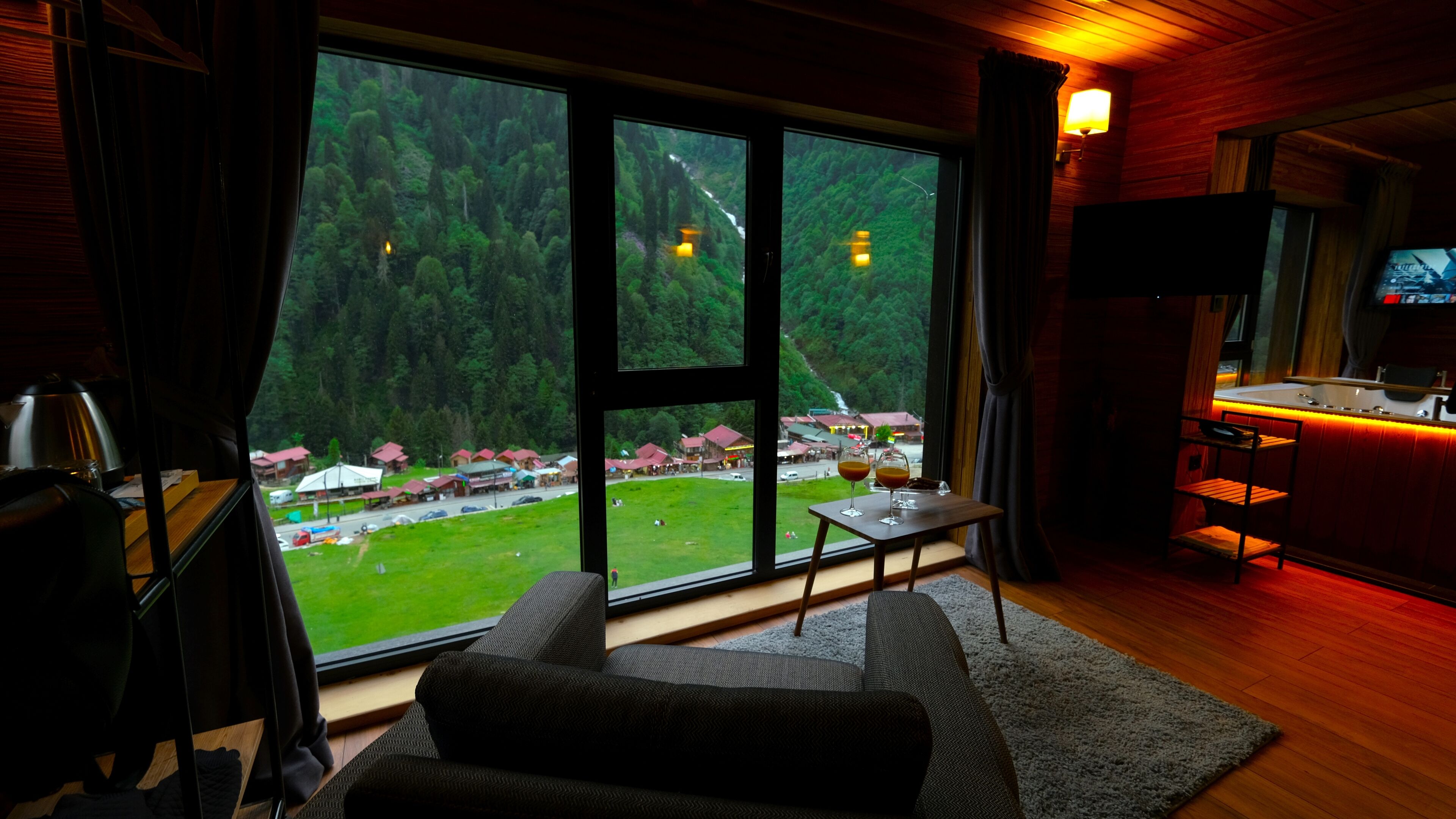 A cozy room with large windows overlooking a forested area, a small table with a drink, and a TV on the right.