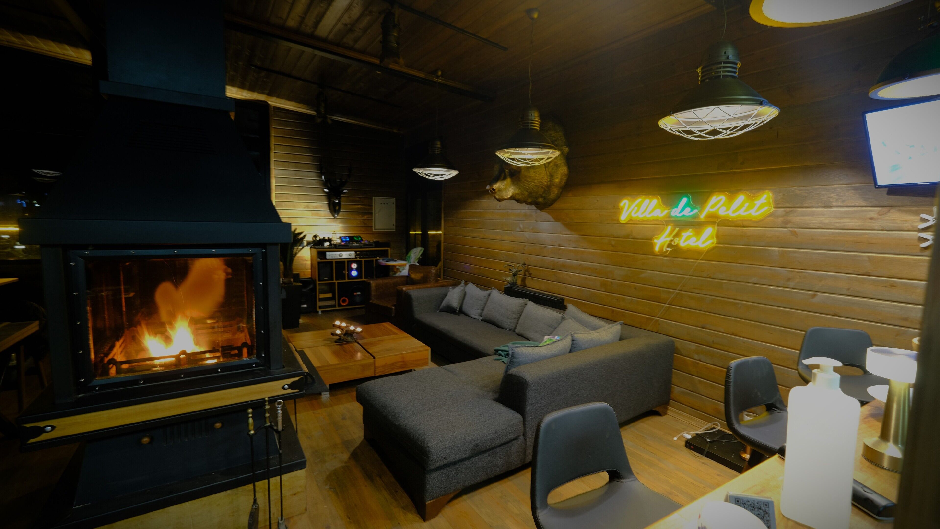 Un salon chaleureux avec une cheminée, un canapé et un panneau lumineux au néon sur lequel on peut lire : « Villa de Pelit Hotel ».