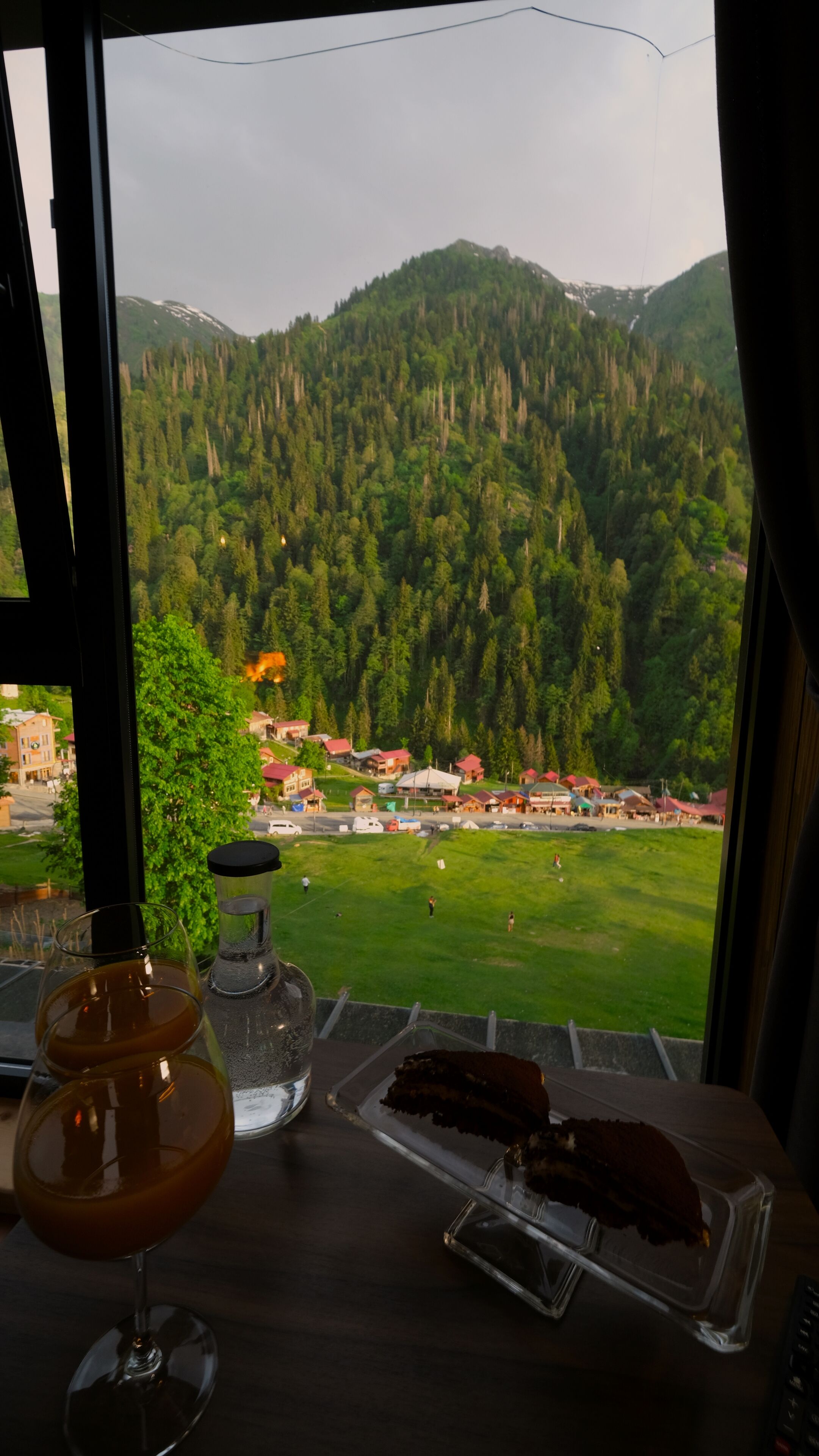 A table with a glass of drink, a jar, and a plate with food, overlooking a mountainous landscape with a village.