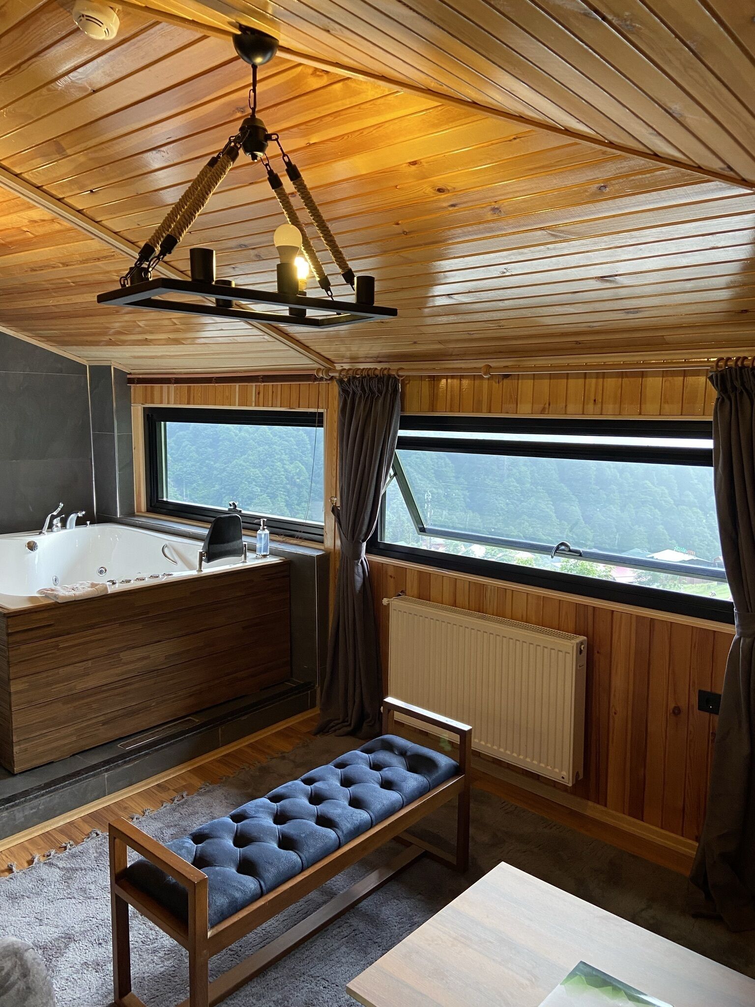 Interior de una cabaña con techo de madera, un ventanal grande, una bañera, un banco y un radiador.