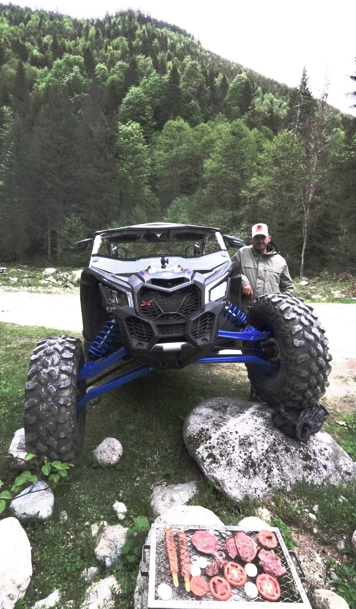 A vehicle with large tires and a rugged design, parked on a grassy area with rocks and a grill with food items.
