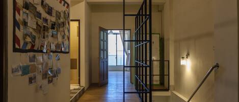 Un pasillo con piso de madera, una pared con fotos colgadas, una escalera con barandilla de metal y una puerta que da a una habitación luminosa.