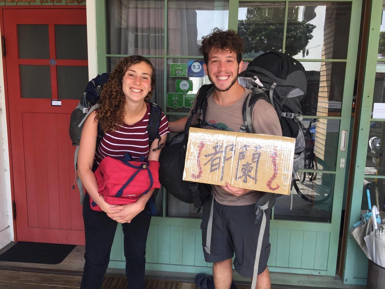Two people standing in front of a door with a red frame, holding a cardboard box with Japanese characters.