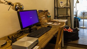 Uma mesa de madeira com um computador, teclado e monitor. Há uma luminária, um ventilador e uma estante com livros e um aviso.