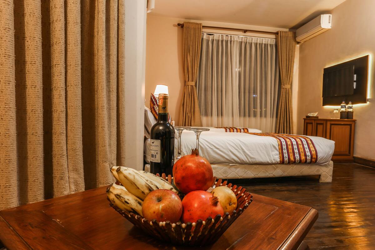 A hotel room with a wooden table, a bed with a striped blanket, a pomegranate, bananas, and a bottle of wine.