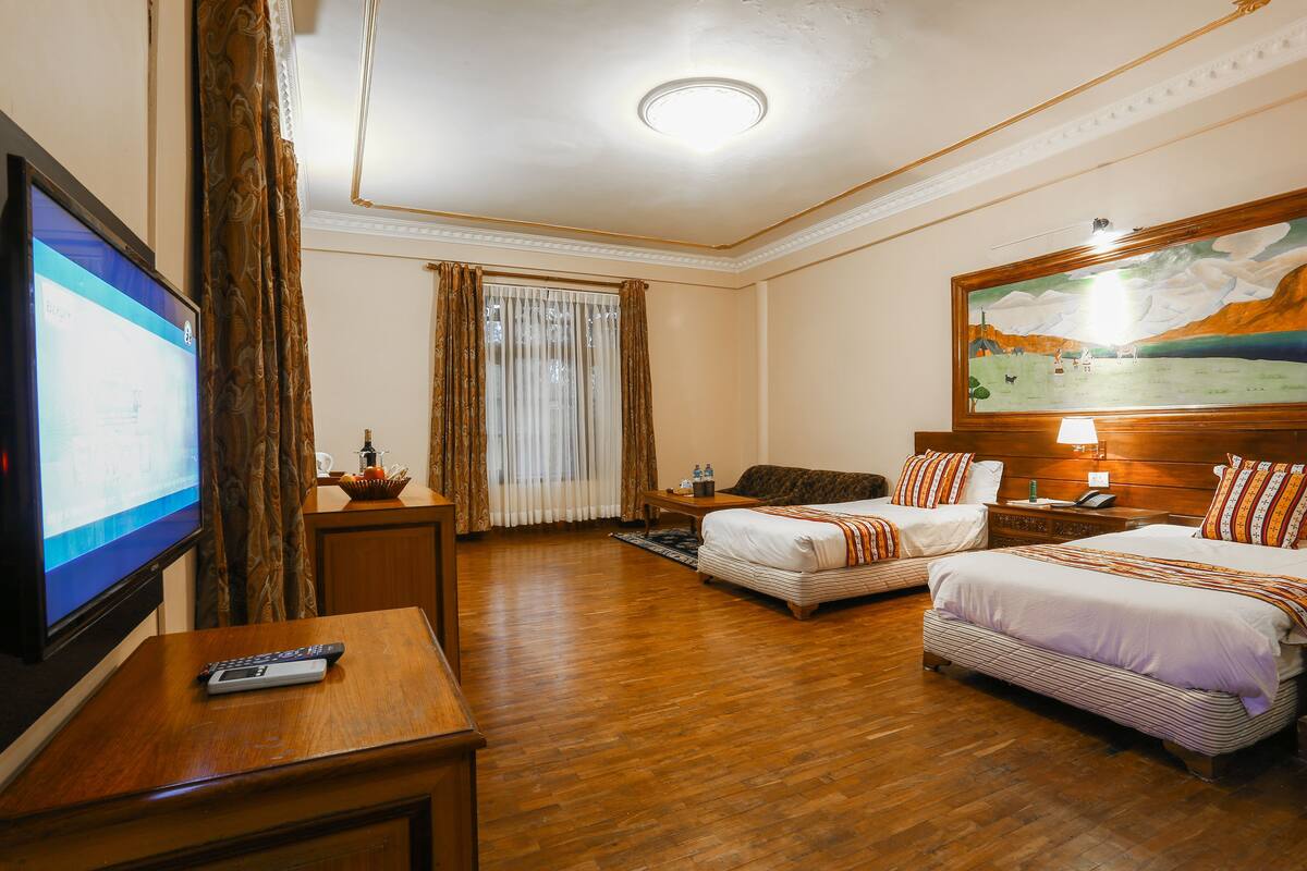 A hotel room with two beds, a television, a remote control, a painting on the wall, and a window with curtains.