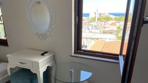 Een slaapkamer met een witte kaptafel, een rond glazen tafeltje, een blauwe stoel en een raam met uitzicht op een stad en zee.