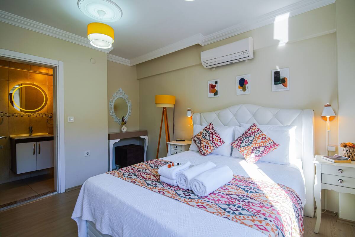 A well-lit bedroom with a large bed, a bedside table, an air conditioning unit, and a mirror on the wall.
