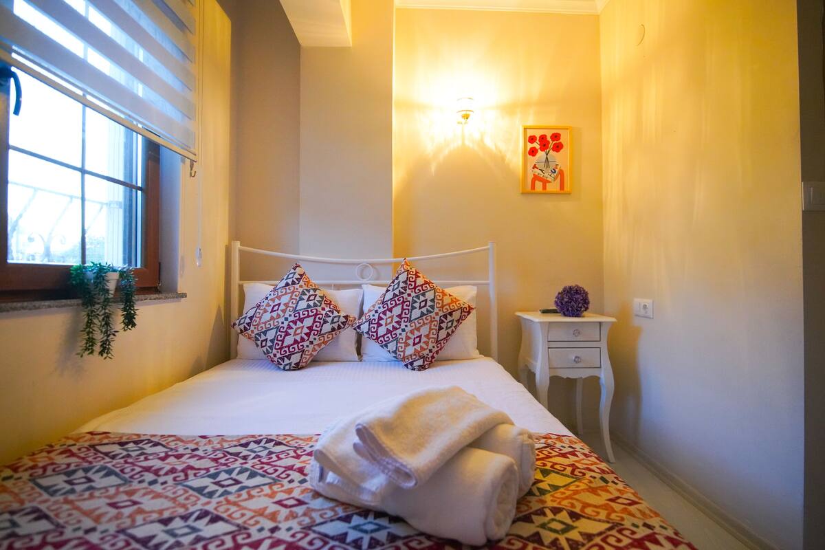 A neatly made bed with patterned bedding, a bedside table with a vase, and a wall-mounted light fixture.