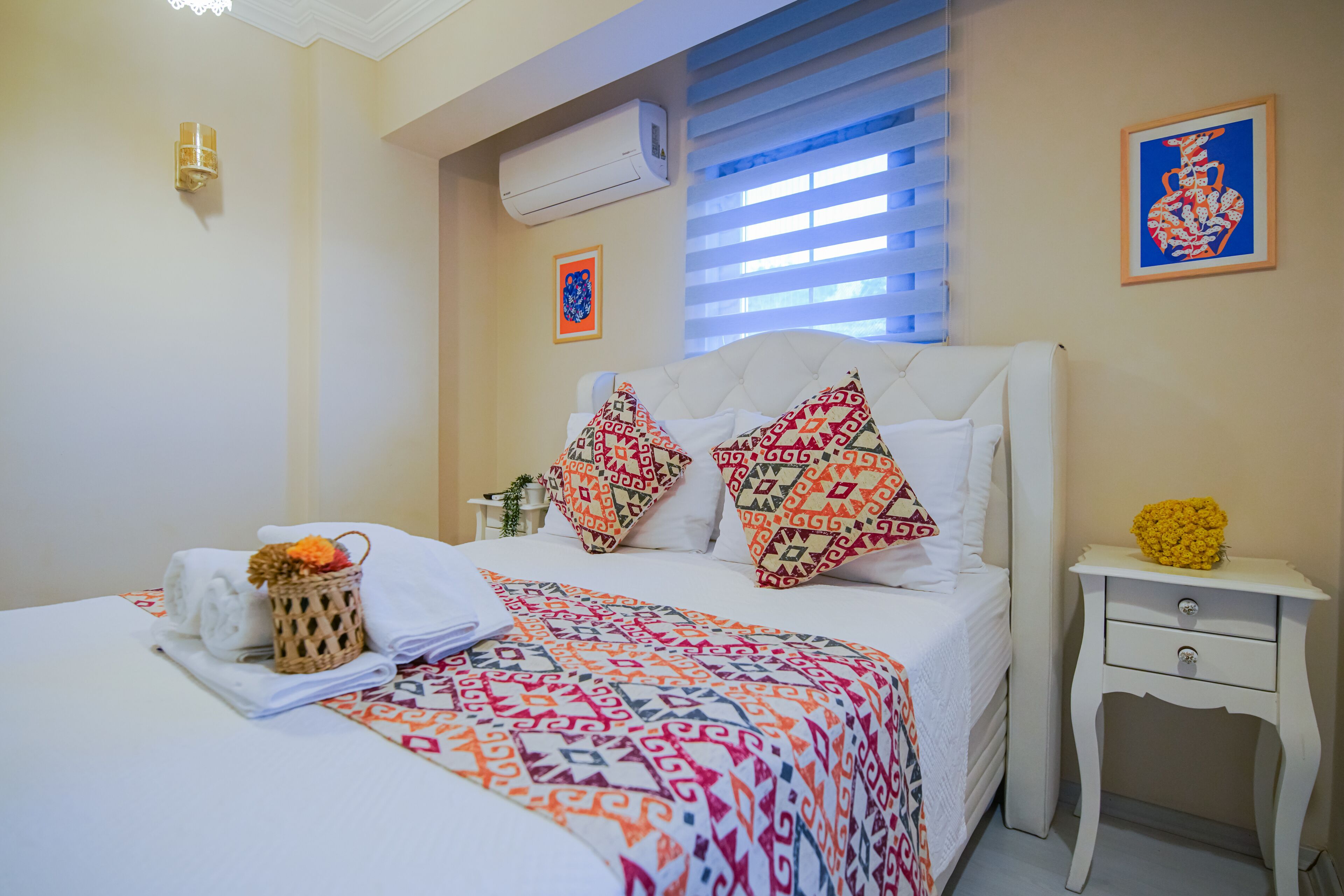 A neatly made bed with patterned pillows, a bedside table with a vase of flowers, and a wall-mounted air conditioner.