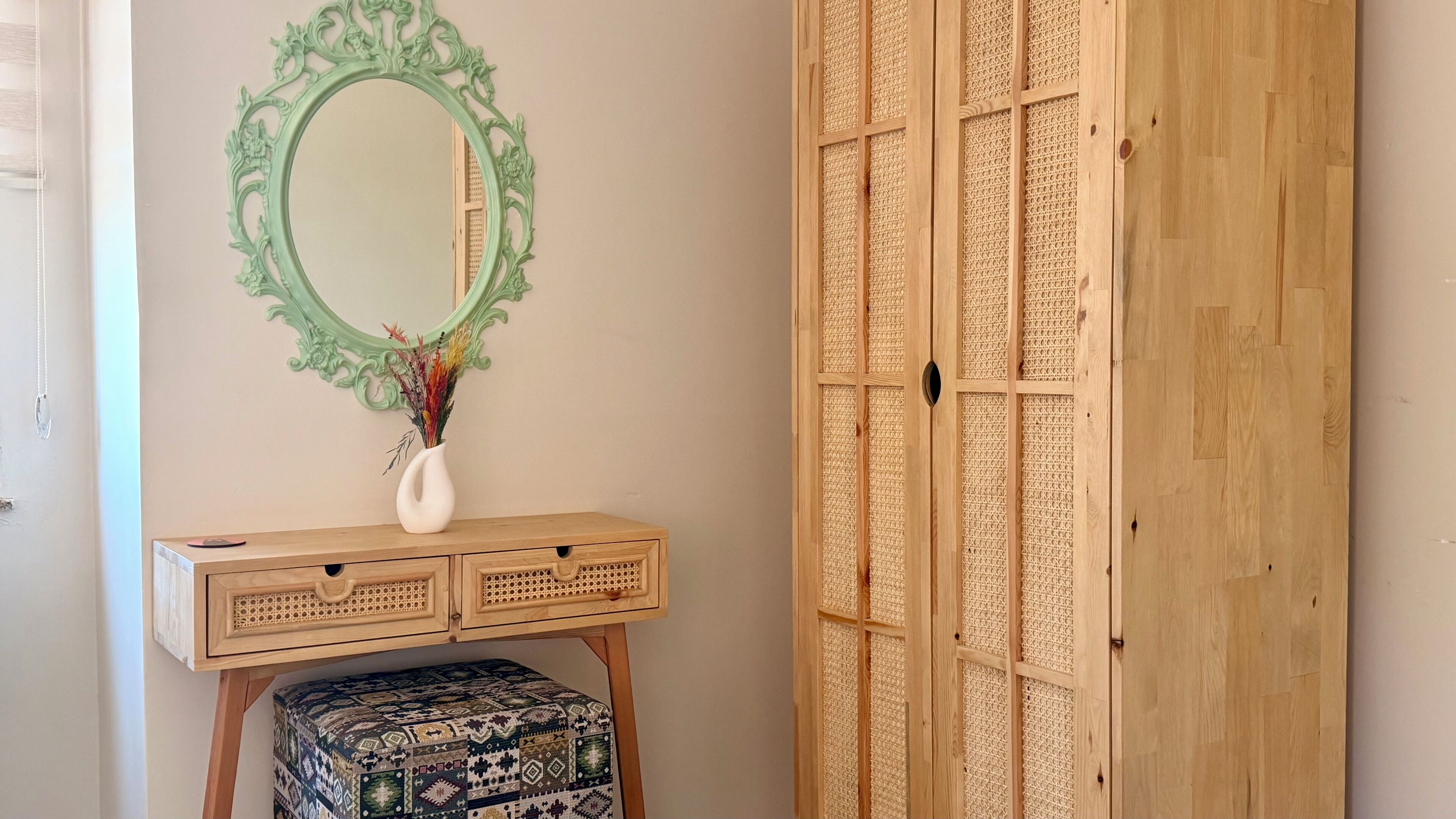 A room with a wooden wardrobe, a mirror with an ornate frame, a wooden dresser with drawers, and a patterned ottoman.