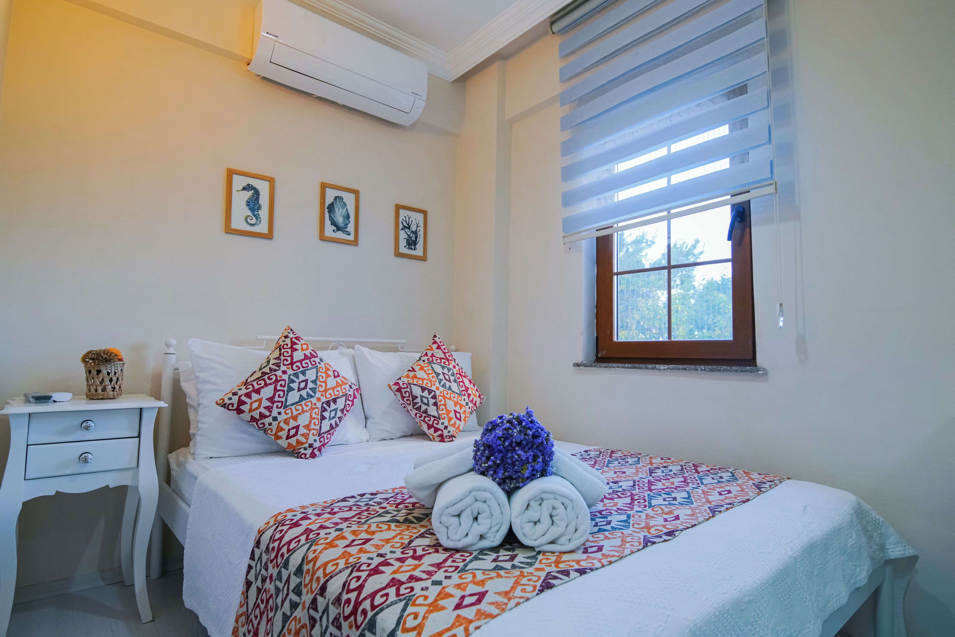 A neatly made bed with patterned bedding, a bedside table with a lamp, and a window with blinds.