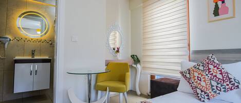 Habitación de hotel con cama, un pequeño escritorio con silla, espejo, ventana con persianas y baño con lavamanos y espejo.