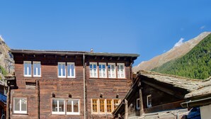 A wooden building with multiple windows and a sloped roof, surrounded by mountains.