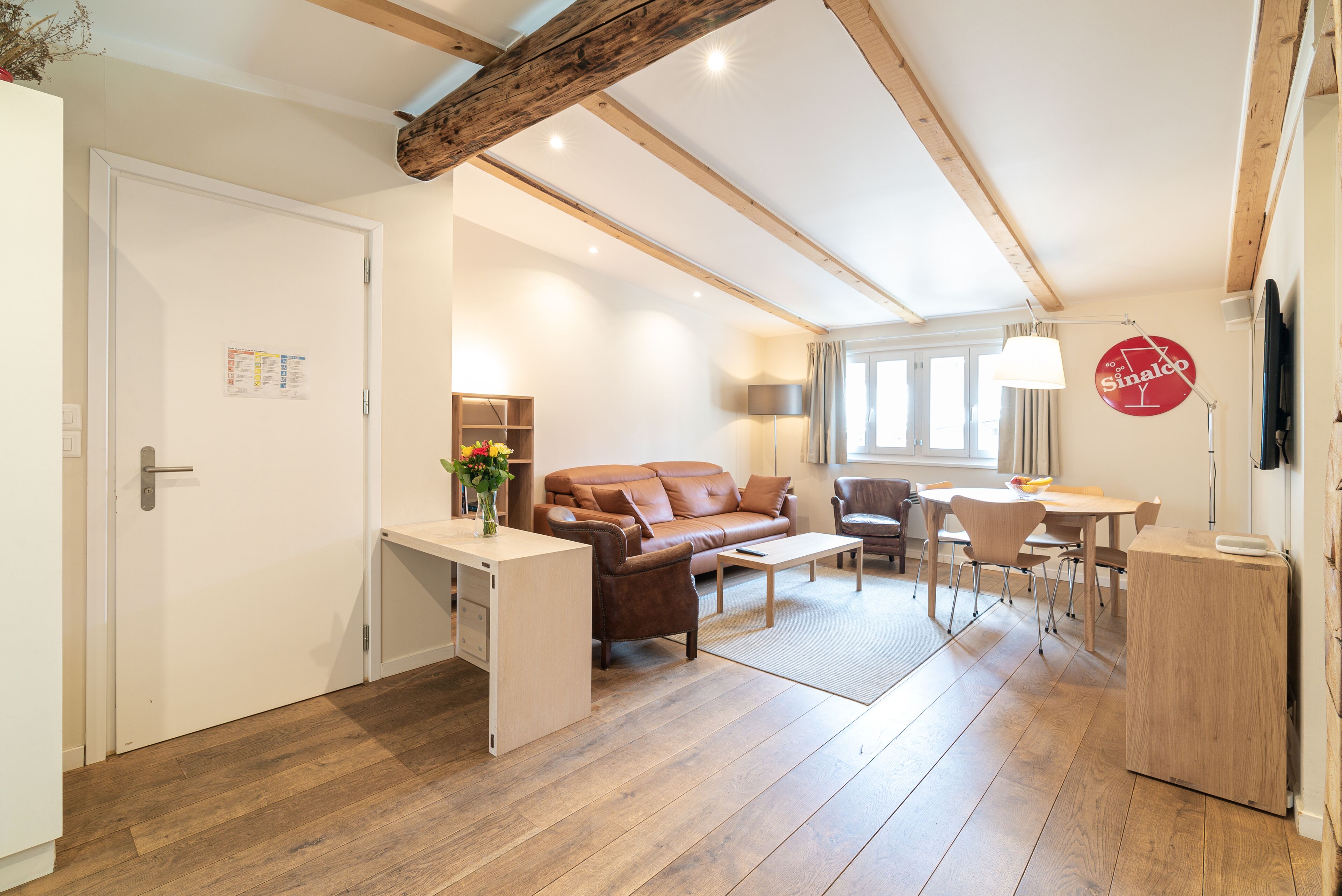 A spacious living area with a wooden floor, a brown sofa, a dining table with chairs, and a small white desk.