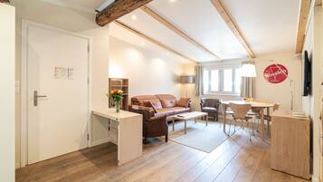 A spacious living area with a wooden floor, a brown sofa, a dining table with chairs, and a small white desk.