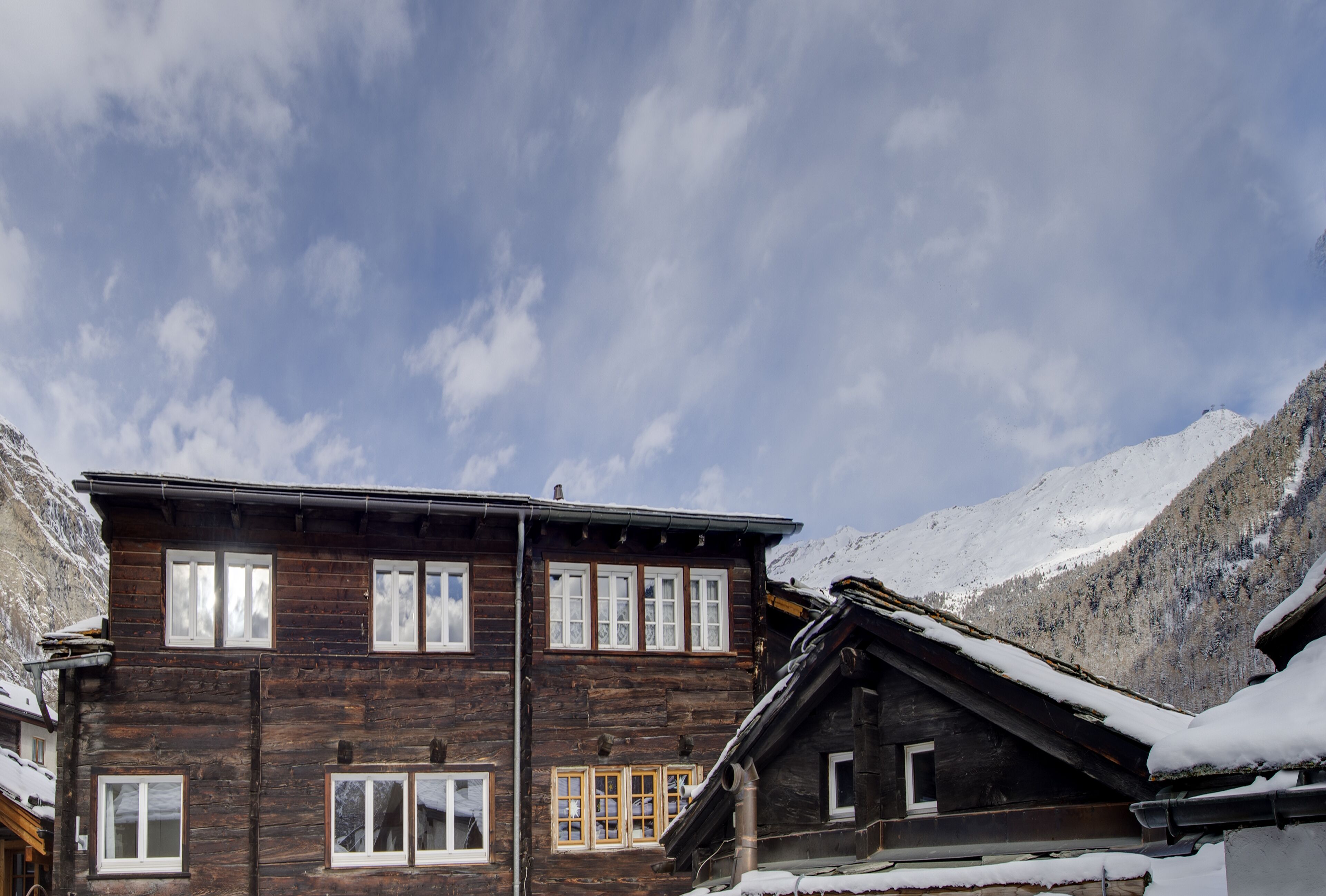 A wooden chalet with multiple windows and a snow-covered roof, surrounded by mountains.