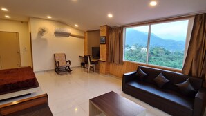 Una habitación de hotel moderna con un ventanal grande que ofrece vistas a la montaña, una zona de estar acogedora con un sofá y una silla, un escritorio con una computadora y un televisor de pantalla plana.