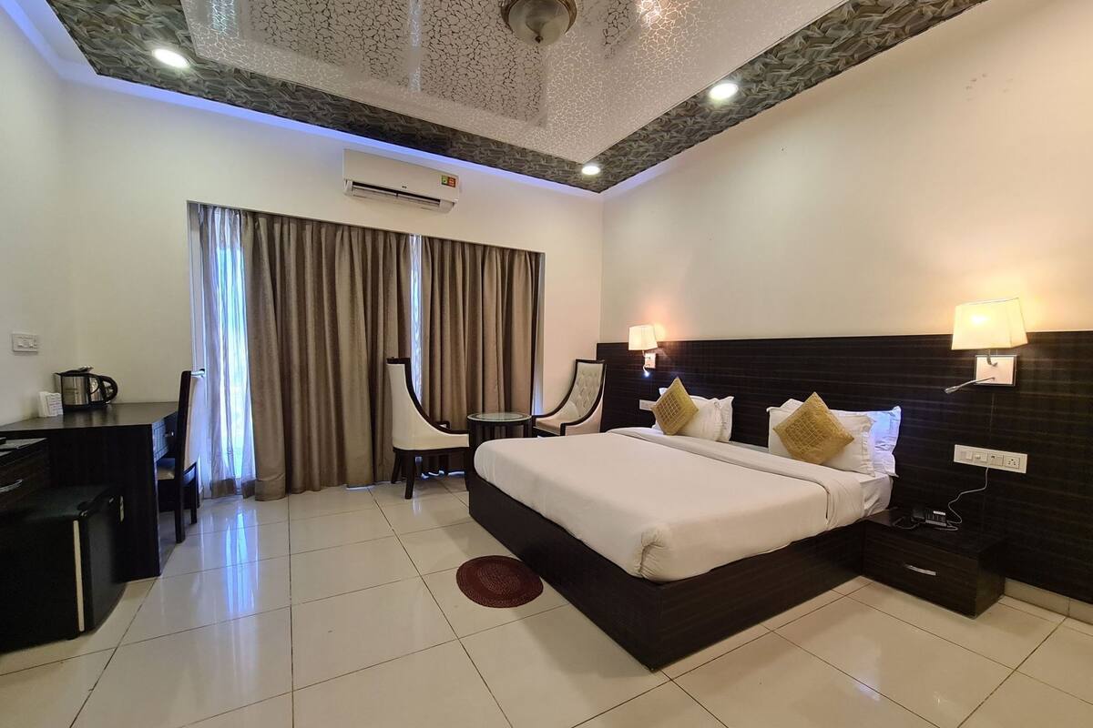 A hotel room with a large bed, a desk with a chair, a small table, and a window with curtains.