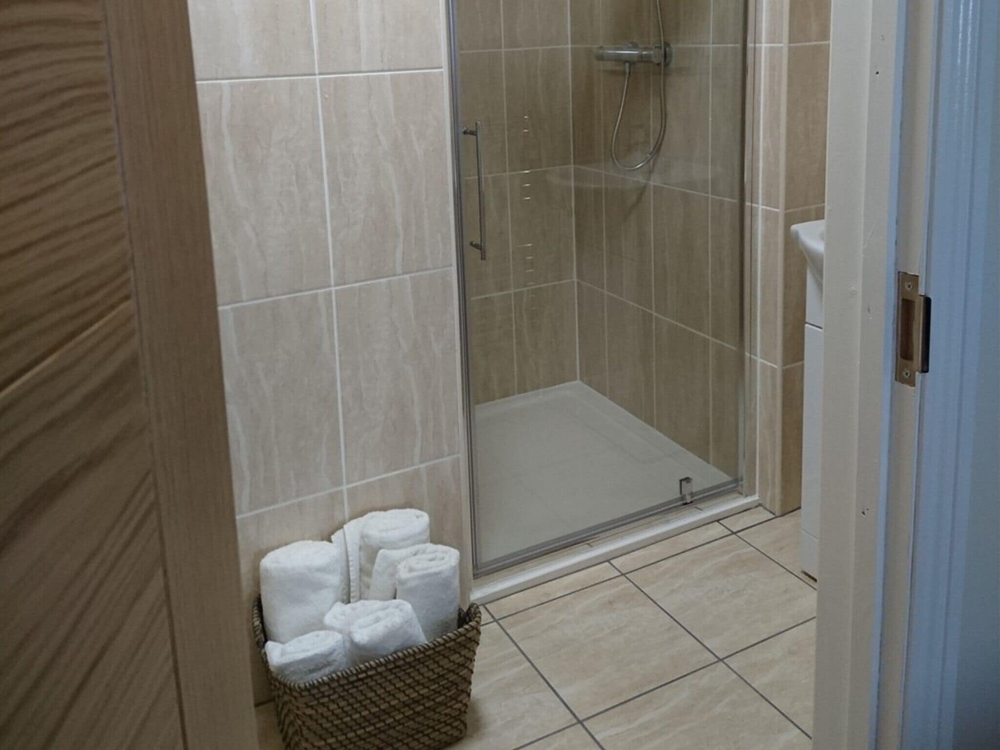 Zona de ducha con puerta de vidrio, un lavamanos blanco y una cesta de mimbre con toallas enrolladas.