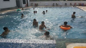A swimming pool with multiple people enjoying the water, surrounded by a white building and some outdoor furniture. - Suva Hotel (Birendranagar)