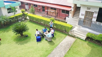 Une cour où un groupe de personnes est assis autour d’une table, entourée de haies bien entretenues et de palmiers.