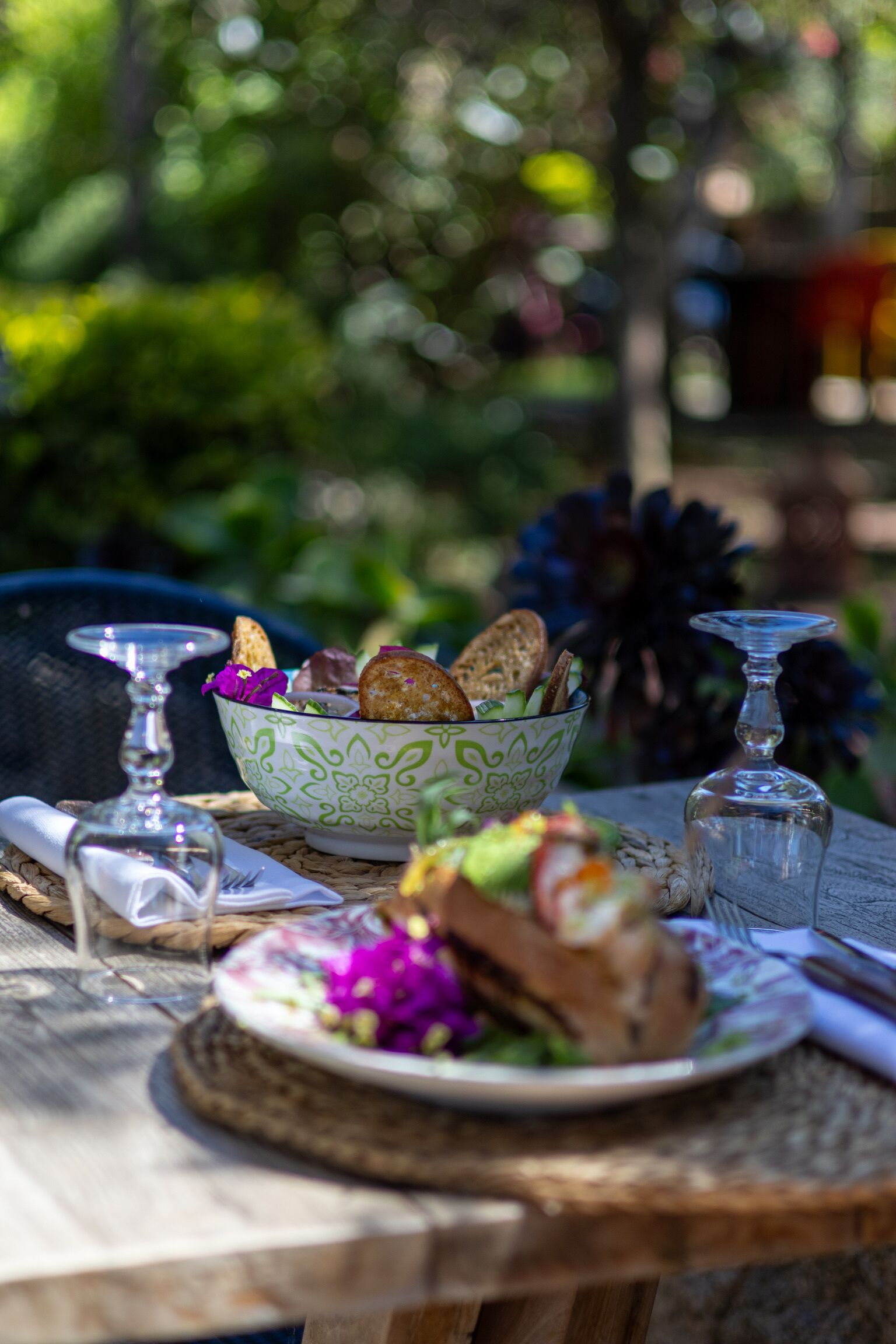 A table set with a bowl of salad, bread, and a glass of water, with a wicker chair and a glass in the background.
