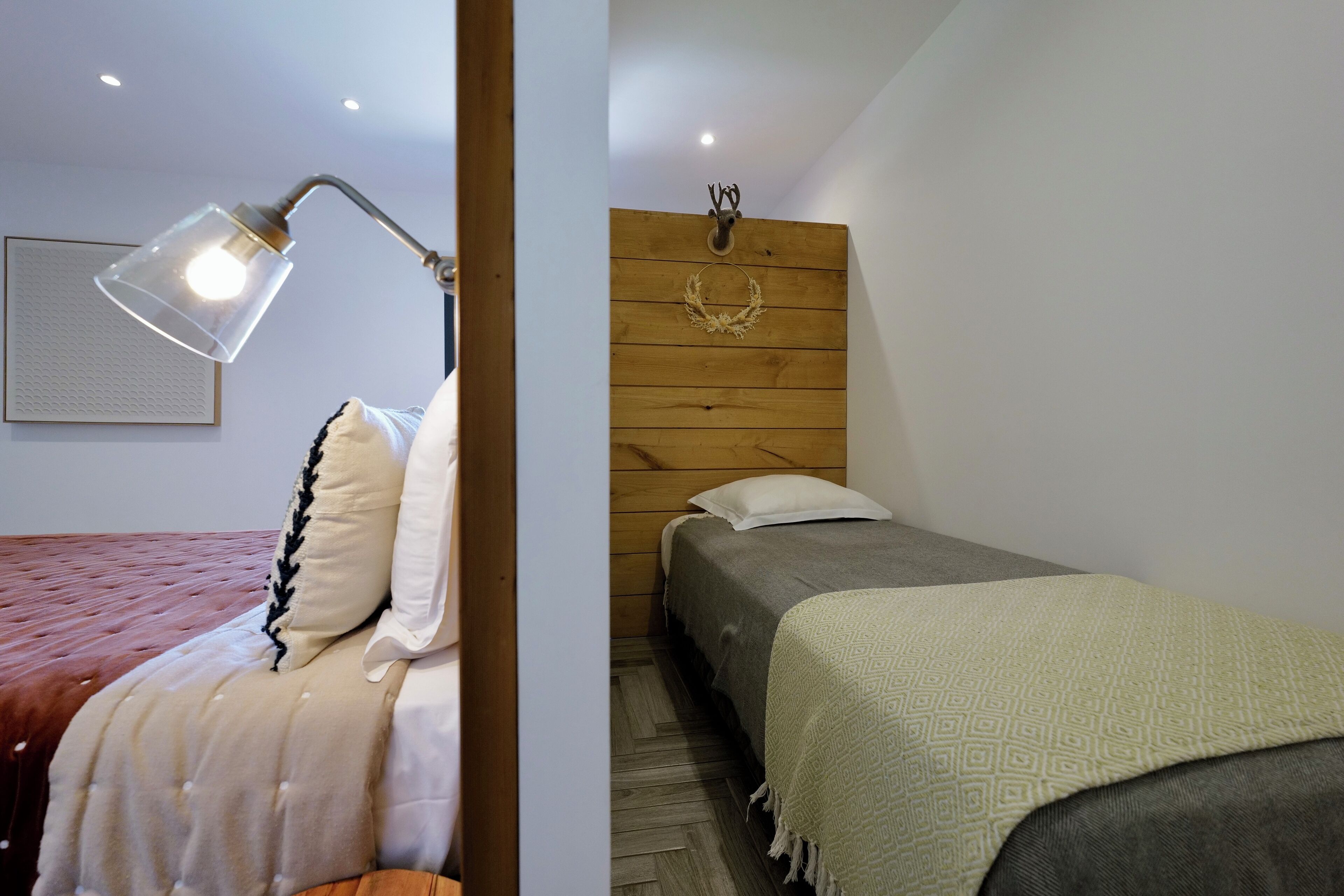 A bedroom with a wooden headboard, a bed with a patterned blanket, a bedside lamp, and a wall-mounted deer head decoration.