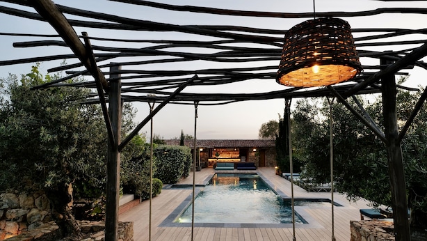 A pool area with a wooden pergola, stone walls, and a hanging light fixture. - Pozzo di Mastri (Figari)